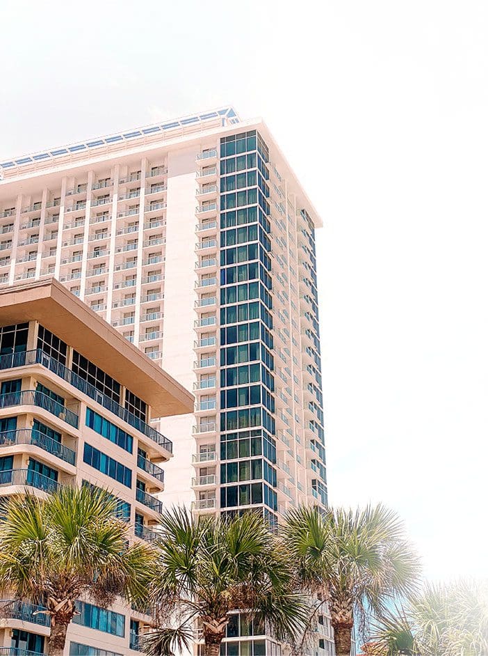 A towering skyscraper stands beside a smaller building, surrounded by lush palm trees